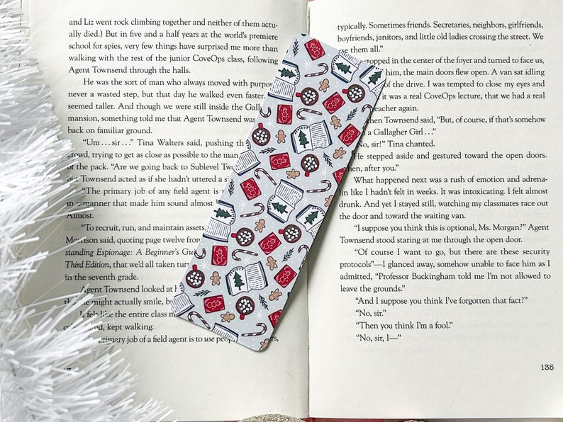 May include: A white and gray bookmark with a festive Christmas pattern. The bookmark features illustrations of gingerbread cookies, candy canes, mugs of hot chocolate, and books. The pattern is repeated throughout the bookmark.