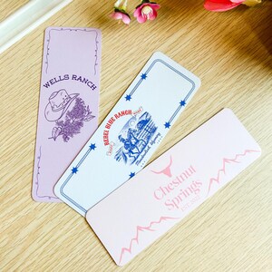 May include: Three rectangular bookmarks with Western-themed designs. One is lavender with a cowboy hat and floral illustration, another is white with a blue illustration of a cowboy on a horse, and the last is pink with text.