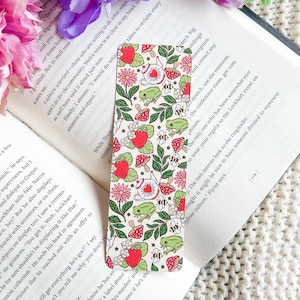 May include: A bookmark featuring a whimsical design with red mushrooms, green frogs, and white teacups on a white background. The bookmark is decorated with a floral pattern and has a white border.