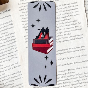 Peut inclure: Marque-page gris avec une pile de livres et une paire d'escarpins noirs à semelles rouges. Des graphiques d'éclats d'étoiles et d'étoiles noirs sont également présents.