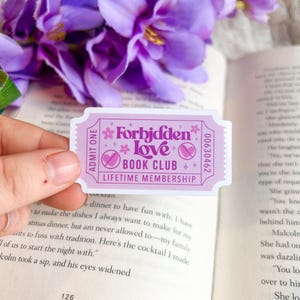 May include: A light purple ticket-shaped sticker with the text "Forbidden Love Book Club Lifetime Membership" in white and pink. The sticker has a floral and heart design. The sticker is held over an open book.