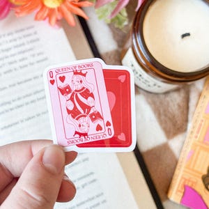 May include: A pink and red sticker featuring a queen of hearts illustration with the text "Queen of Books".