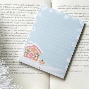 May include: A light blue notepad with a winter theme. The notepad features a gingerbread house illustration with pink and white accents, snowflakes, and lined pages. The notepad is approximately 10 cm x 15 cm.