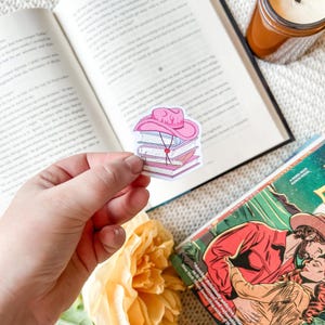 May include: A pink cowboy hat sticker atop a stack of books, held in a hand. The sticker has a red heart detail and a string. The background includes an open book, a candle, and a book cover with the text "Another deliciously addictive romas".