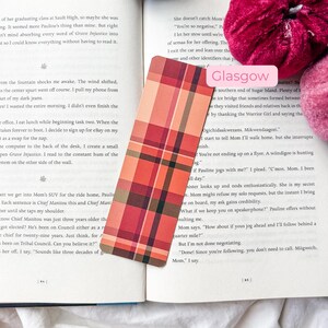 K&ouml;nnte beinhalten: Ein rechteckiger Lesezeichen mit einem Karomuster in Rot-, Orange- und Braunt&ouml;nen. Das Lesezeichen liegt auf einem offenen Buch mit Text. Ein rosa Etikett mit dem Wort "Glasgow" ist sichtbar.
