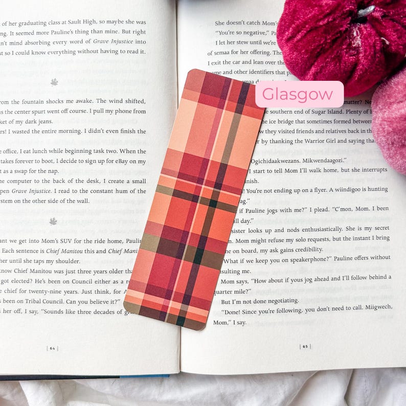 K&ouml;nnte beinhalten: Ein rechteckiger Lesezeichen mit einem Karomuster in Rot-, Orange- und Braunt&ouml;nen. Das Lesezeichen liegt auf einem offenen Buch mit Text. Ein rosa Etikett mit dem Wort "Glasgow" ist sichtbar.