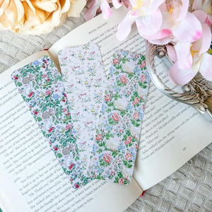 May include: Three decorative bookmarks with floral patterns. One displays rabbits and flowers, another has a repeating pattern of small white cats with bows, and the third features roses and other flowers. They are on an open book.