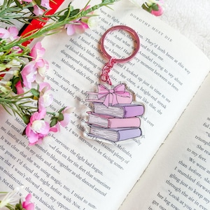 May include: A pink keyring with a design of stacked books and a bow. The books are in shades of pink and purple. The keyring is on an open book with text and pink flowers.