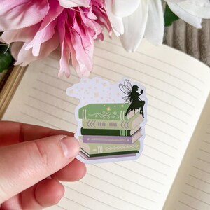 May include: A sticker featuring a silhouette of a fairy sitting on a stack of books. The books are green, purple, and brown with white accents. The fairy has wings and is holding a book. The sticker is on a white background.