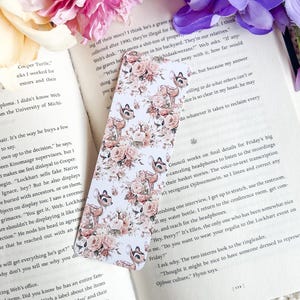 May include: A white bookmark with a floral pattern featuring pink roses, green leaves, and brown deer silhouettes. The bookmark is designed with a vintage aesthetic.