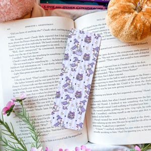May include: A bookmark with a repeating pattern of cartoon ducks in witch hats, holding wands and brewing potions, with gift boxes and crescent moons on a lavender striped background. Ideal for fantasy readers.