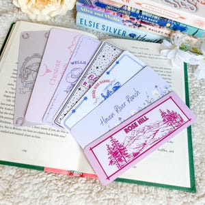 May include: A selection of rectangular bookmarks with different designs and text, such as "Gold R", "Chestnut", "Wells", "Rebel Blue Ranch", "Haven River Ranch", and "Rose Hill". The bookmarks are in pink, lavender, and white, on an open book.