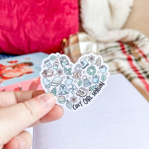 May include: A heart-shaped sticker with winter-themed illustrations, including a candle, mittens, and a gingerbread house. The sticker has the text "Cozy Girl Season" in a handwritten font.
