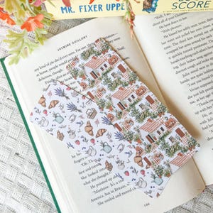 May include: Two decorative bookmarks with charming illustrations. One features a pattern of houses, while the other showcases croissants, lavender, teapots, and strawberries. The bookmarks are designed for use in books and are ideal for readers.