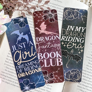 May include: Four colourful bookmarks with dragon designs and text. The bookmarks feature a dark blue bookmark with the text "Just a Girl Dreaming of Riding Dragons", a dark red bookmark with the text "Dragon Fantasy Book Club", a dark blue bookmark with the text "In My Dragon Riding Era", and a dark blue bookmark with the text "Just a Girl Dreaming of Riding Dragons".