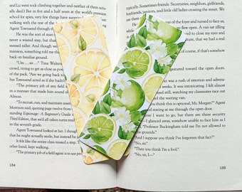 May include: Two bookmarks with a watercolor design of lemons and limes. The bookmarks are white with green leaves and white flowers. The text on the book reads "The primary job of a field agent is to use people typically. Sometimes friends. Secretaries, neighbors, girlfriends, boyfriends, janitors, and little old ladies crossing the street. We use them..."