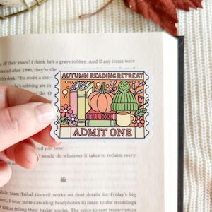 May include: A rectangular sticker with a scalloped edge. It features the text "AUTUMN READING RETREAT" and "ADMIT ONE." The design includes a pumpkin, books, a green beanie, a mug, and flowers. The words "FALL BOOKS" are also present.