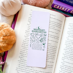 May include: White bookmark featuring the text "Midnight Margaritas" and green illustrations of a cocktail, coffee pot, and snake. The bookmark is on an open book.