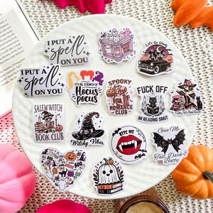 May include: A collection of Halloween themed stickers featuring various designs, including a witch's hat, a vampire's mouth, a ghost, a book, and a bat. The stickers are on a white surface with a pink and orange bat in the background.