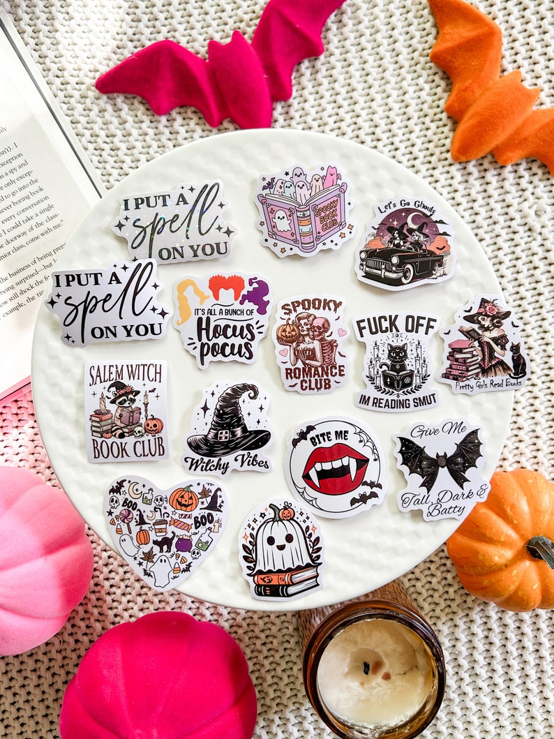 May include: A collection of Halloween themed stickers featuring various designs, including a witch's hat, a vampire's mouth, a ghost, a book, and a bat. The stickers are on a white surface with a pink and orange bat in the background.