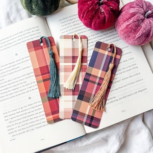 K&ouml;nnte beinhalten: Drei Lesezeichen mit Karomuster und farbigen Quasten liegen auf einem offenen Buch. Die Lesezeichen weisen rot-, orange- und violett karierte Designs auf. Dekorative Samtk&uuml;rbisse in Gr&uuml;n- und Burgundert&ouml;nen befinden sich im Hintergrund.