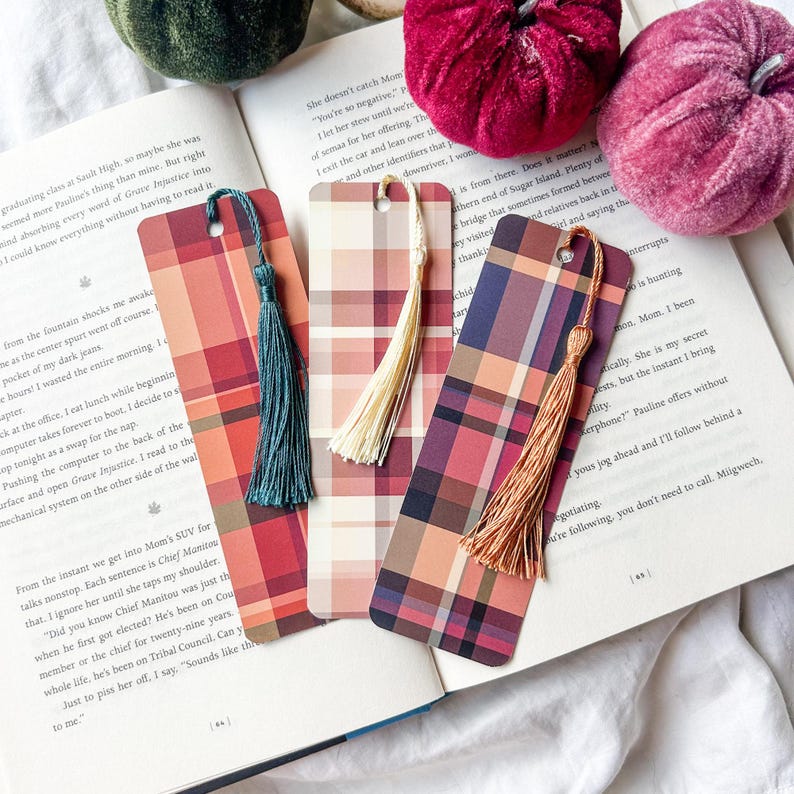 K&ouml;nnte beinhalten: Drei Lesezeichen mit Karomuster und farbigen Quasten liegen auf einem offenen Buch. Die Lesezeichen weisen rot-, orange- und violett karierte Designs auf. Dekorative Samtk&uuml;rbisse in Gr&uuml;n- und Burgundert&ouml;nen befinden sich im Hintergrund.