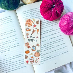 May include: A white bookmark with autumn-themed illustrations and the text "Hello Autumn." The design includes pumpkins, leaves, a sweater, and a coffee cup. The bookmark is lying on an open book.