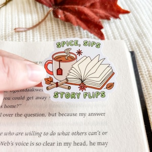May include: A sticker with the text "SPICE, SIPS STORY FLIPS" depicts a mug of tea, an open book, and cinnamon sticks. The sticker is held over an open book with text.