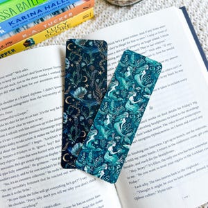 Peut inclure: Deux marque-pages décoratifs avec des motifs célestes et de sirènes. L'un présente un fond bleu foncé avec des lunes, des étoiles et des coquillages. L'autre a un fond turquoise avec des sirènes. Les deux sont rectangulaires et posés sur un livre ouvert.