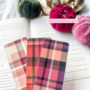 K&ouml;nnte beinhalten: Drei Lesezeichen mit Karomuster in Rosa-, Rot- und Oranget&ouml;nen. Die Lesezeichen liegen vor einem offenen Buch und Samtk&uuml;rbissen. Der Text "Lamination Example" ist sichtbar.