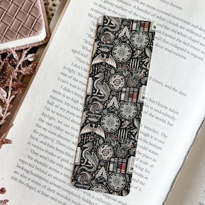 May include: A black and white bookmark featuring a detailed design of a dragon, a compass, and other mystical symbols. The bookmark is printed with a repeating pattern and has a slightly textured surface.
