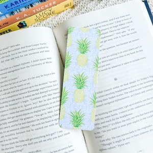 May include: A rectangular bookmark featuring a light blue backdrop and a repeating pattern of yellow pineapples with green leaves. The bookmark is inserted into an open book.