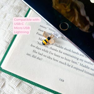 May include: A gold-coloured bee charm with black and yellow stripes and rhinestone accents. The charm is attached to a clear phone accessory. Text on the image reads "Compatible with: USB-C, Micro USB, Lightning."
