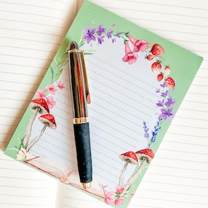 Peut inclure: Un bloc-notes vert avec une bordure florale représentant des champignons, des fraises et des fleurs. Un stylo noir est posé sur le bloc-notes.