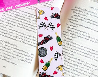 May include: A pink bookmark with racing-themed illustrations. It features red race cars, checkered flags, champagne bottles, gold trophies, and red hearts. The bookmark is set against the backdrop of an open book.