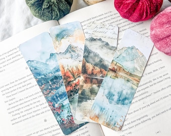 May include: Four watercolour landscape bookmarks depicting mountains, lakes, and forests. The bookmarks are rectangular with rounded corners and feature blues, greens, and oranges. They are placed on an open book.