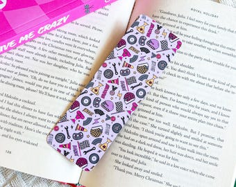 May include: A light purple bookmark with a racing theme, featuring illustrations of helmets, wrenches, checkered flags, and trophies. The bookmark is placed in an open book with text on the pages.