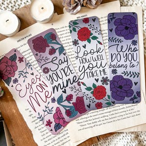 May include: Four floral bookmarks with different quotes. The bookmarks are purple, pink, and gray with white lettering. The quotes are "Eyes on Me", "Say You're Mine", "Look How Well You Take Me", and "Who Do You Belong To?"