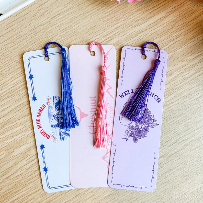 May include: Three rectangular bookmarks with decorative tassels. The bookmarks are white, pink, and lavender, with text and illustrations. The white bookmark has blue accents and text that reads "Blue Ranch". The pink bookmark says "Chestnut". The lavender bookmark says "Wells Ranch".