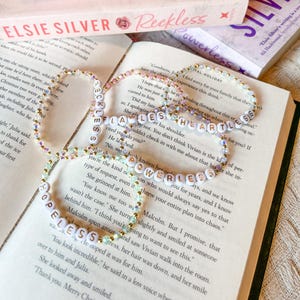 May include: Three beaded bracelets with white letter beads spelling "Reckless", "Powerless", and "Hopeless" are displayed on an open book. The bracelets are accented with gold and iridescent beads. The book is open to a page of printed text.