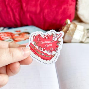 May include: A heart-shaped sticker with a red cake design, featuring white frosting, candy canes, and a snowman. The sticker has the words "Christmas Girlie" in the centre. The sticker is held over an open notebook.