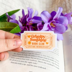 May include: A sticker with the text "Grumpy Sunshine Book Club Lifetime Membership" in orange on a cream background. The sticker has a ticket shape with hearts and a sun graphic. Purple flowers and a book are in the background.