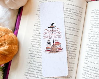 May include: A white bookmark with the text "MY BROOMSTICK RUNS ON BOOKS & LATTES" features a witch's hat, broom, coffee cups, and stacks of books. The bookmark is set on an open book.
