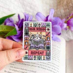 May include: A rectangular sticker with a Halloween theme. The sticker features the words "READ.SCREAM" and "HORROR.REPEAT" with illustrations of ghosts, pumpkins, and other spooky elements. The sticker is held in a hand.