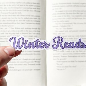 May include: A white sticker with the text "Winter Reads" in purple cursive lettering. The sticker is being held in a hand over an open book.