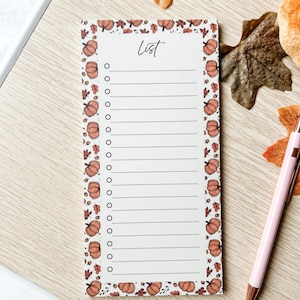 Peut inclure: Bloc-notes rectangulaire sur le thème de l'automne. Le bloc-notes a un fond blanc avec une bordure de citrouilles et de feuilles orange. Le mot "List" est écrit en haut. Un stylo rose est posé sur le côté.