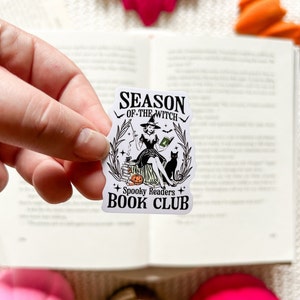 May include: A white sticker with a black and white illustration of a witch sitting on a pumpkin with a black cat. The text on the sticker reads "Season of the Witch Spooky Readers Book Club".