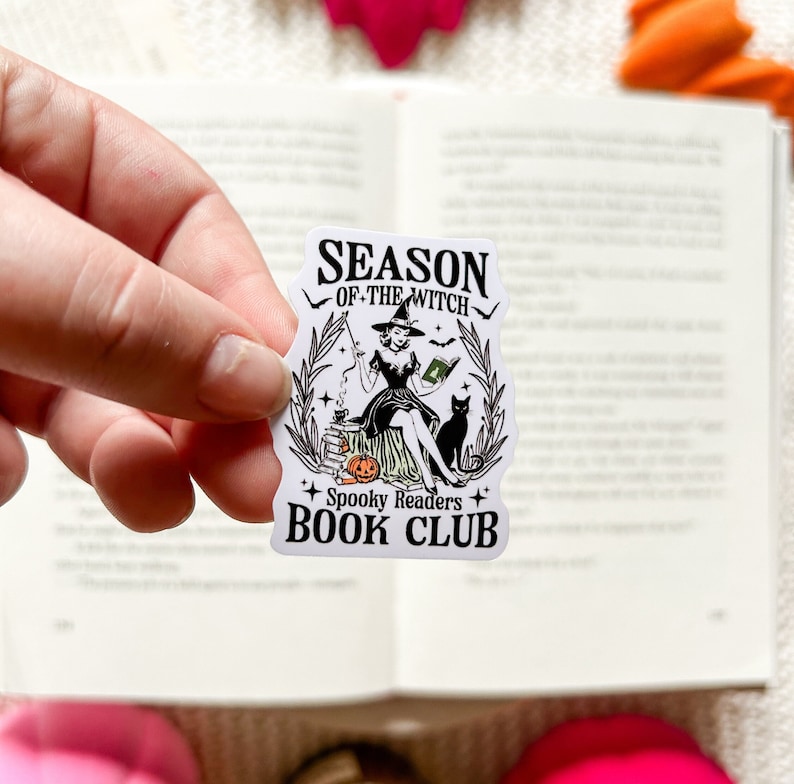 May include: A white sticker with a black and white illustration of a witch sitting on a pumpkin with a black cat. The text on the sticker reads "Season of the Witch Spooky Readers Book Club".