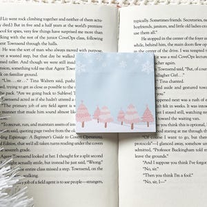 May include: A blue sticky note with a winter scene of five small pink and white trees on a snowy background. The trees are in a row with a white path in front of them. The note is on top of an open book.