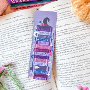 May include: A purple bookmark with a stack of illustrated books, including "Potions" and "Spells." A witch's hat and flowers decorate the top. The bookmark is placed beside an open book.